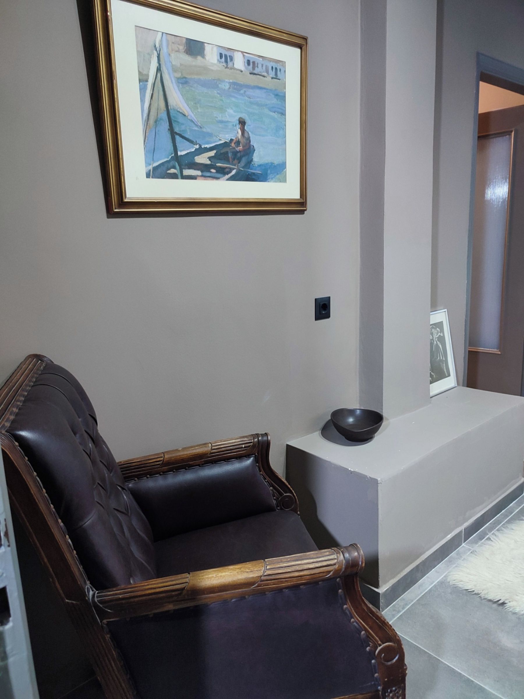 A living room with a dark leather armchair, a framed painting of a sailboat, a small black bowl, and a white rug.