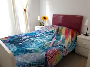 A bed with a colorful, abstract bedspread, a white nightstand with a lamp, and a window with curtains. - Flat With Pool Views in Corralejo (La Oliva)