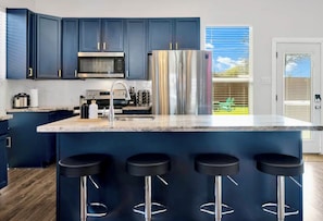 A modern kitchen with dark blue cabinets, stainless steel appliances, a marble countertop, and four bar stools. - Family & Friends Retreat, Hot Tub + Arcade + Cinema (Dallas)