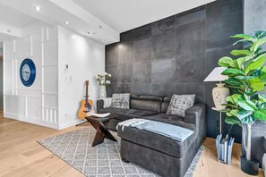 A modern living room with a grey sectional sofa, a patterned rug, a guitar, and a large potted plant. - 🏙️ Penthouse-Style Furnished Condo | Gastown, Downtown Vancouver 🏙️ (Vancouver)