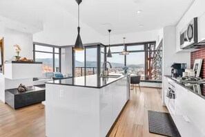 Une cuisine moderne dotée d’un grand îlot central, de luminaires suspendus noirs et offrant une vue sur la ville grâce à de grandes fenêtres.