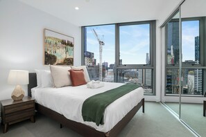 A modern hotel room with a large bed, a nightstand, a lamp, a painting, and a view of the cityscape through large windows. - Modern Southbank Escape | Marco Melbourne (Southbank)