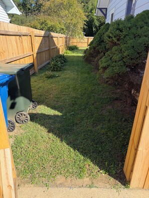 A backyard with a wooden fence, a blue trash bin, and a grassy area. - Charming Blue Cottage - Walking Distance to Downtown (Waynesville)