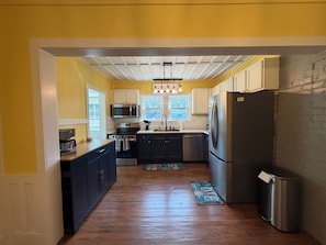 A kitchen with a wooden floor, white cabinets, stainless steel appliances, and a hanging light fixture. - Charming Blue Cottage - Walking Distance to Downtown (Waynesville)