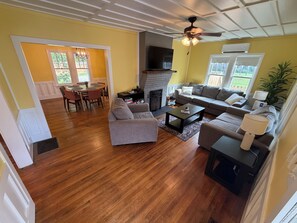 A living room with a fireplace, a ceiling fan, and a dining area with a table and chairs. - Charming Blue Cottage - Walking Distance to Downtown (Waynesville)