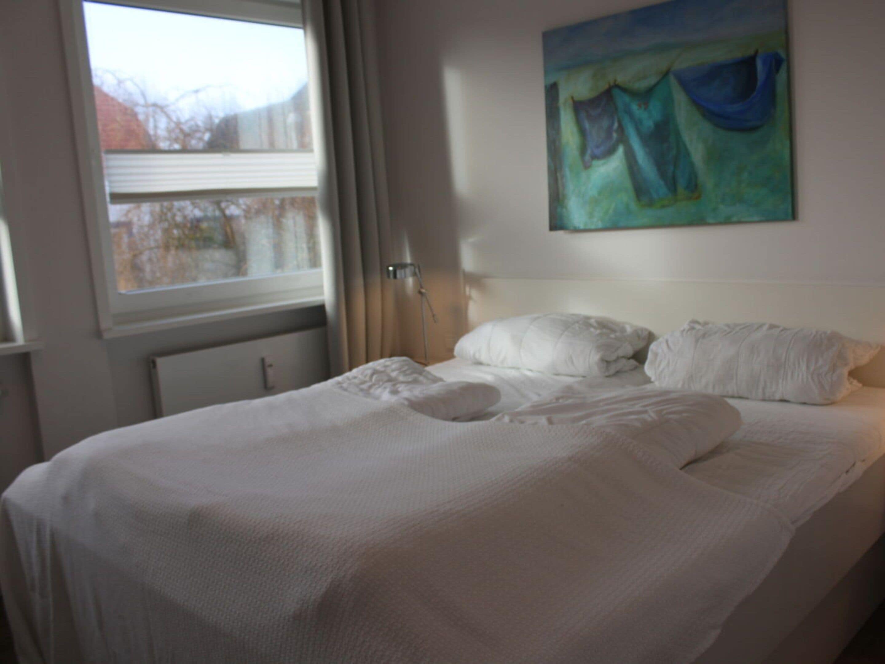 A neatly made bed with white linens in a room with a window and a painting on the wall.