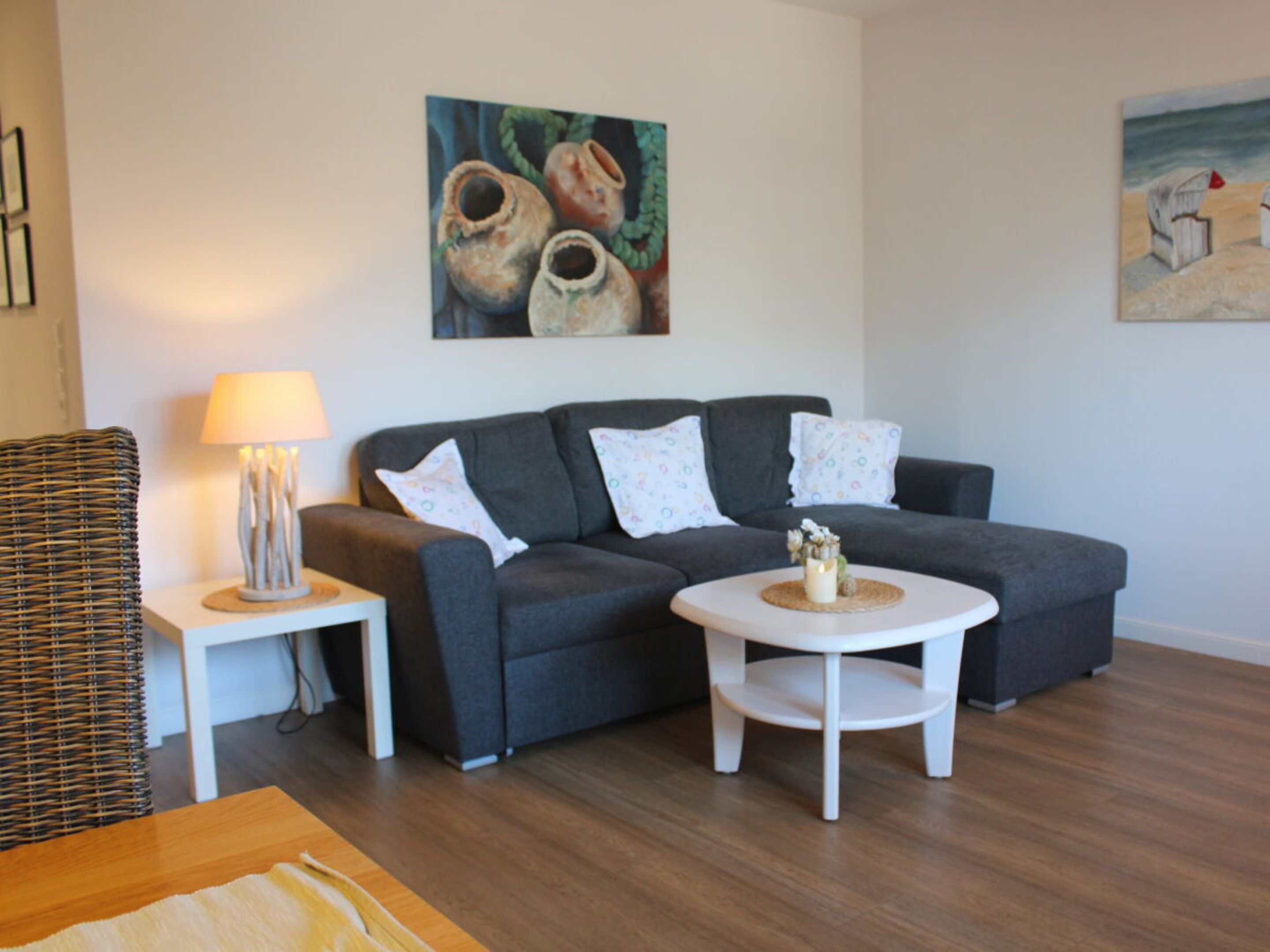 Een woonkamer met een grijze bank, witte kussens, een klein wit tafeltje met een kaars en een rieten stoel.
