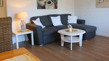 Een woonkamer met een grijze bank, witte kussens, een klein wit tafeltje met een kaars en een rieten stoel.