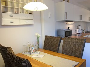 Ein Essbereich mit Korbstühlen, einem Holztisch, einer weißen Lampe und einer Küche im Hintergrund.
