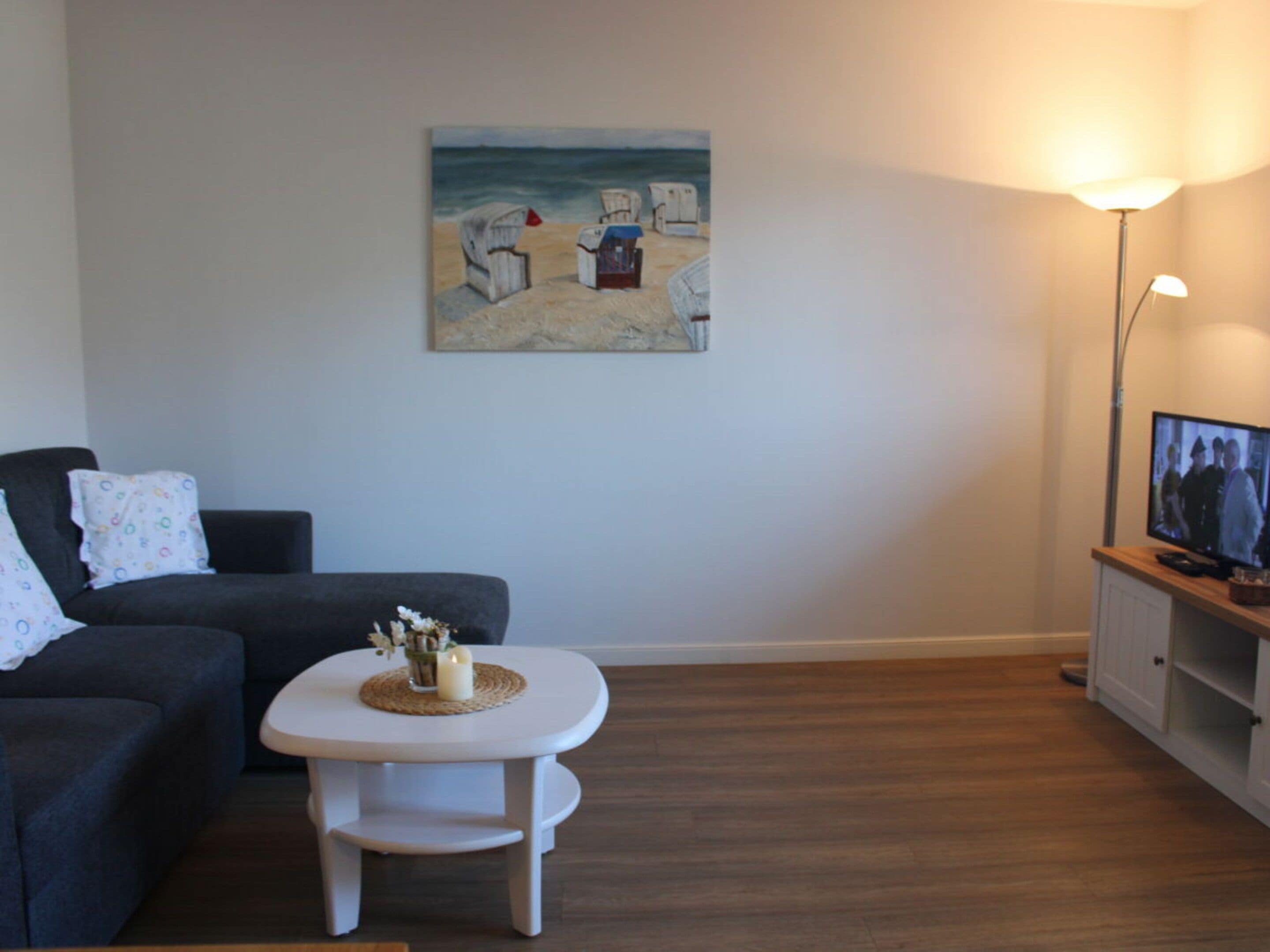 Un salon avec un canapé de couleur sombre, une petite table blanche ornée d’une bougie et de fleurs, un tableau représentant une scène de plage et un téléviseur posé sur un meuble en bois.