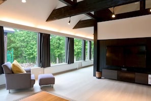 Ein modernes Wohnzimmer mit großem Fernseher, bequemen Sitzgelegenheiten und großen Fenstern mit Blick auf Grünflächen.