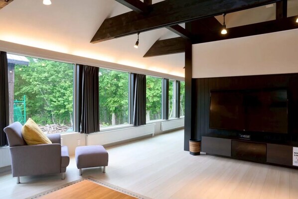 Un salon moderne doté d’un grand téléviseur, de sièges confortables et de grandes fenêtres offrant une vue sur la verdure.