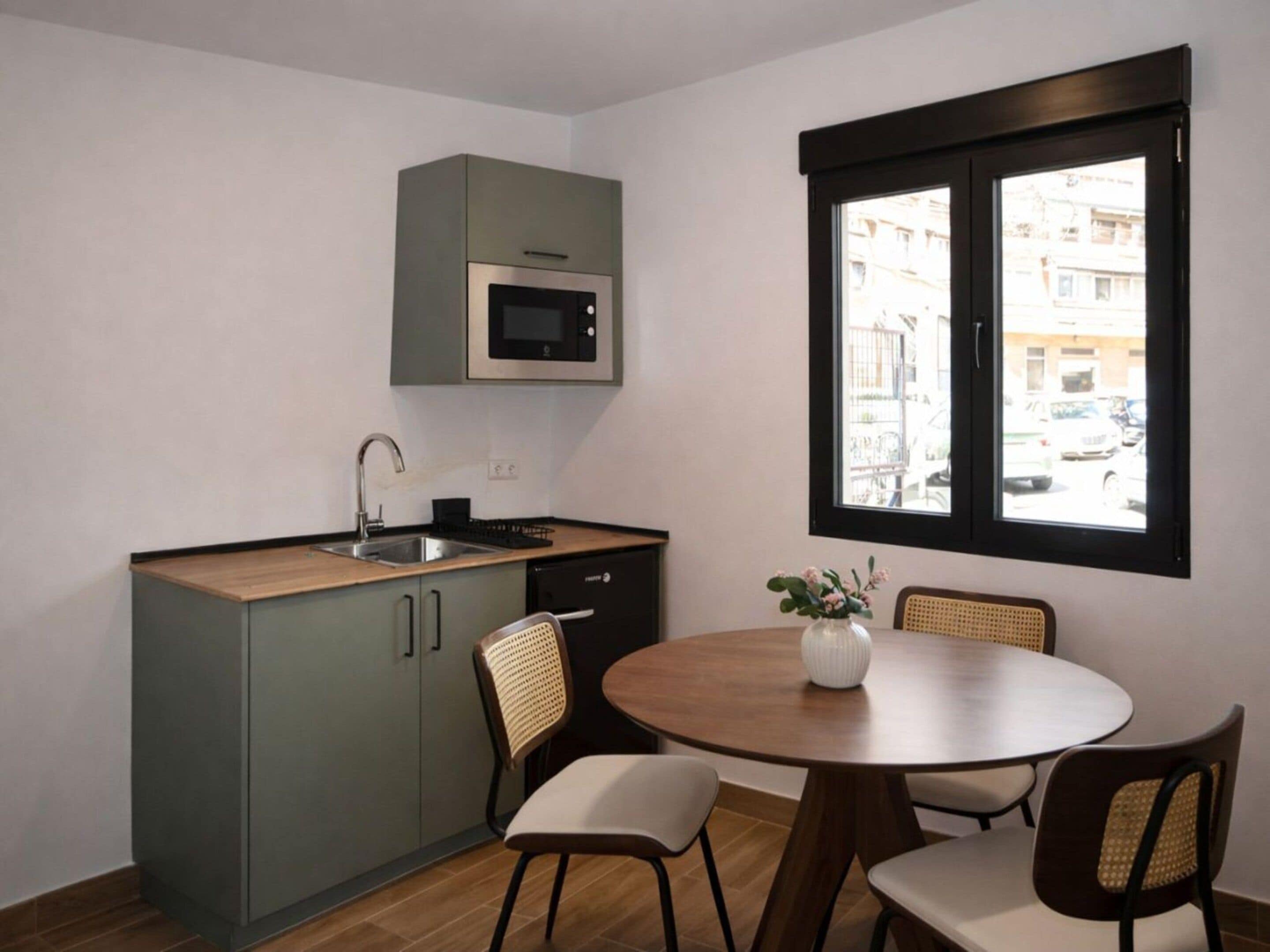Een compacte keuken met een ingebouwde magnetron, een spoelbak en een vaatwasser. Er staat een ronde houten tafel met vier stoelen en een vaas met bloemen.