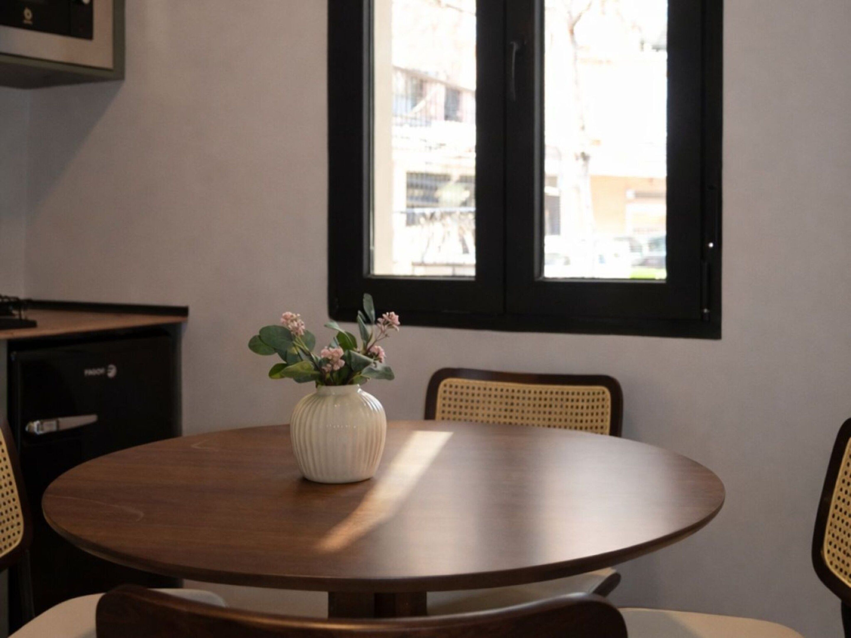 Una mesa redonda de madera con un jarrón blanco que contiene flores rosas, dos sillas de mimbre y una ventana con vistas a edificios.