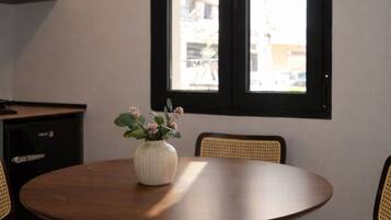 Una mesa redonda de madera con un jarrón blanco que contiene flores rosas, dos sillas de mimbre y una ventana con vistas a edificios.