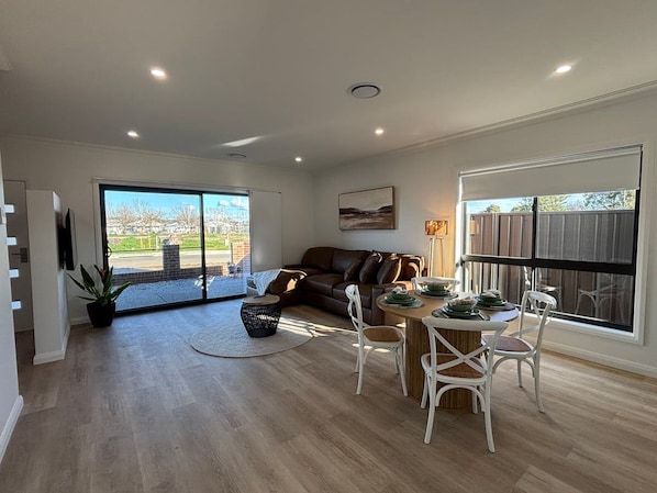 A spacious living area with a sliding glass door leading to a patio, a brown leather sofa, a dining table with white chairs, and a large window with a view of a residential area. - Summons Fourteen - Sleeps 8 (Wagga Wagga)
