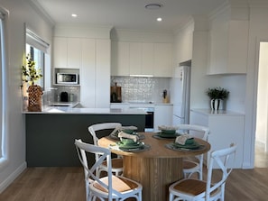 A modern kitchen with white cabinetry, a central island with seating, and a dining area with white chairs. - Summons Fourteen - Sleeps 8 (Wagga Wagga)