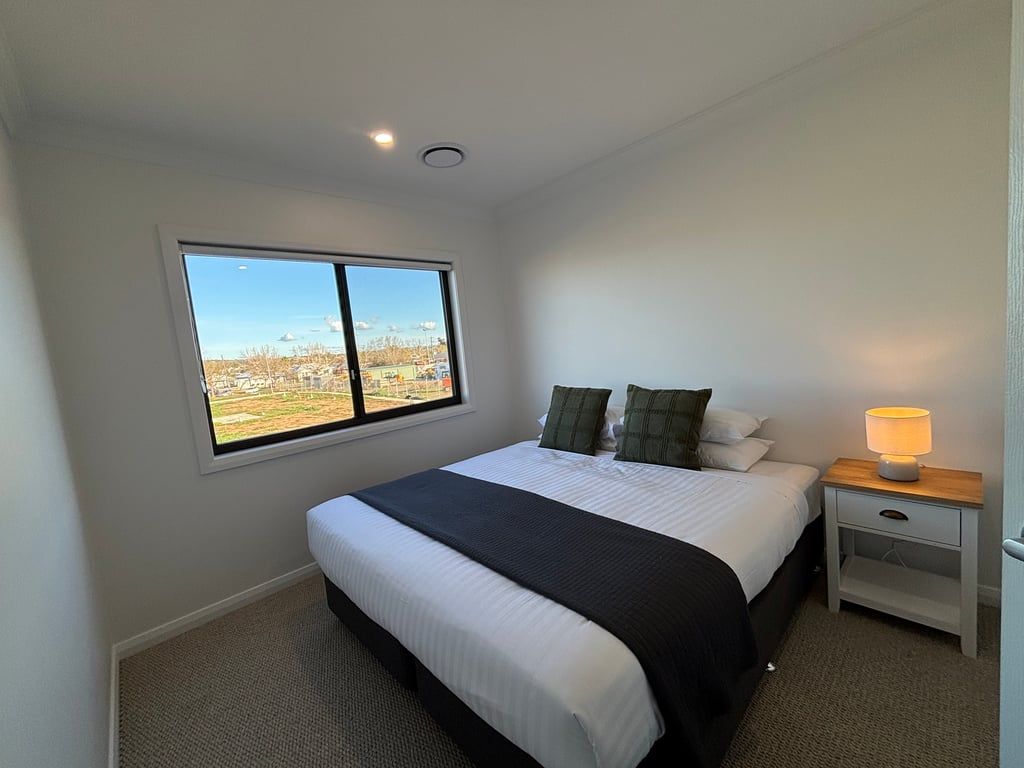 A bedroom with a large window, a bed, a bedside table with a lamp, and a carpeted floor.