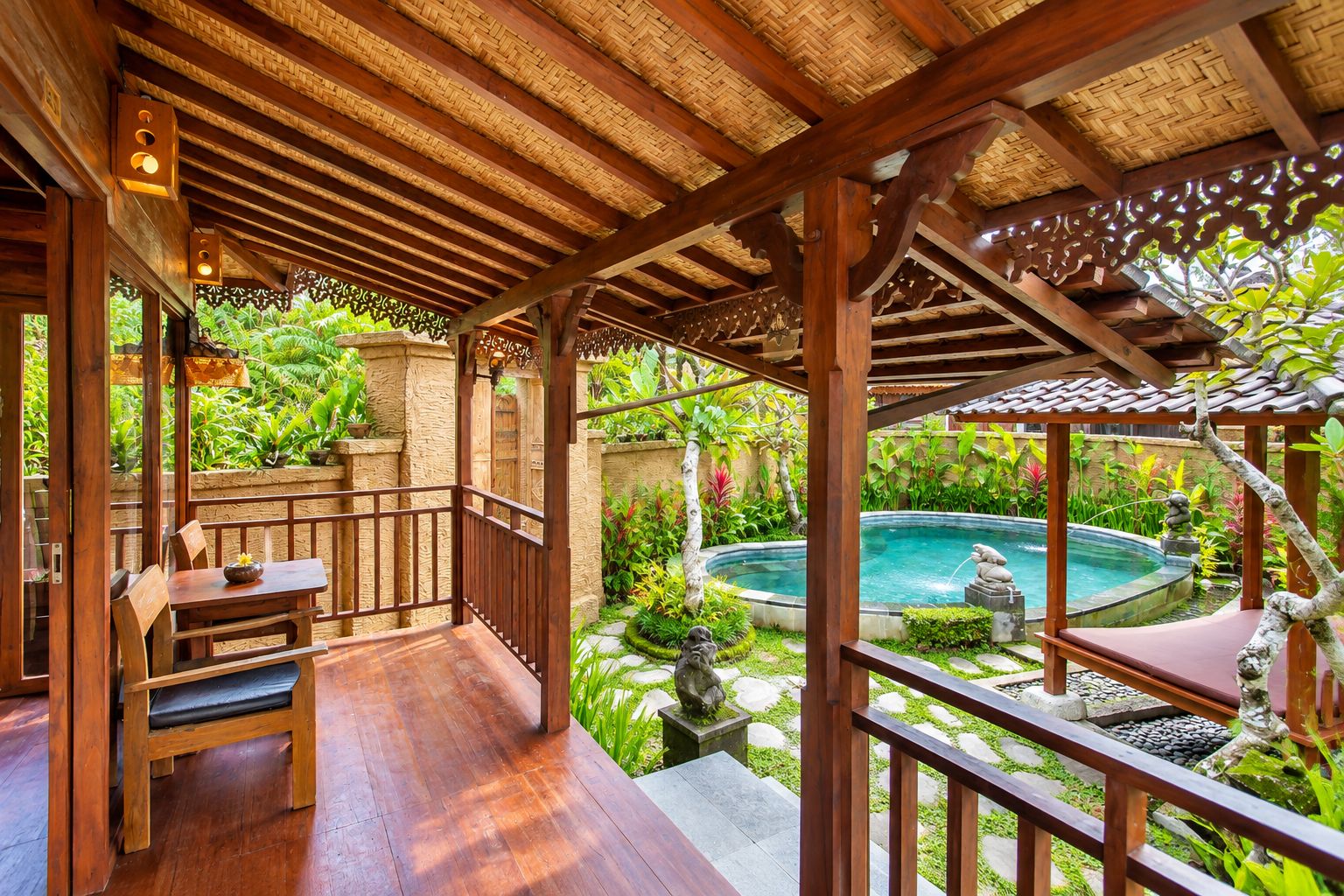 Una terraza de madera con piscina, rodeada de exuberante vegetación y estatuas de piedra.