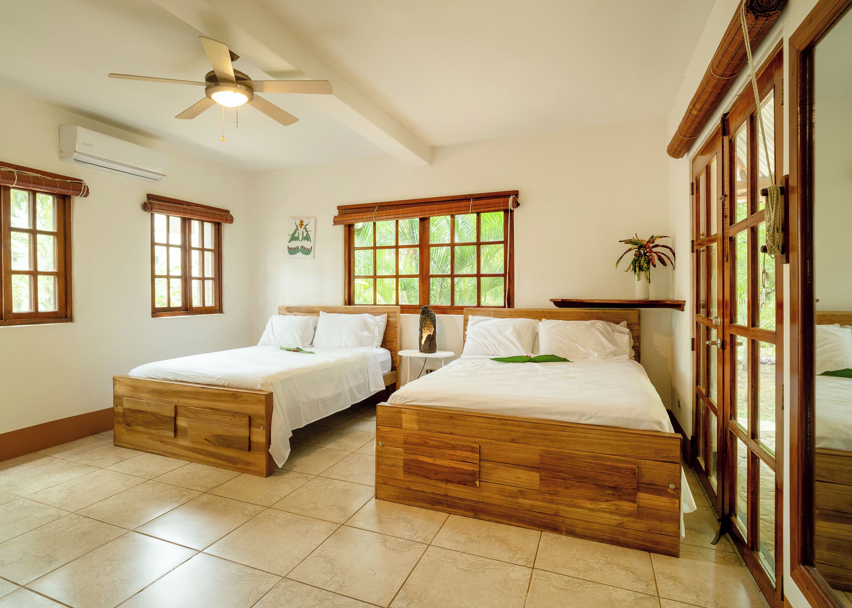 Una habitación con dos camas de madera, un ventilador de techo y ventanas amplias con marcos de madera.