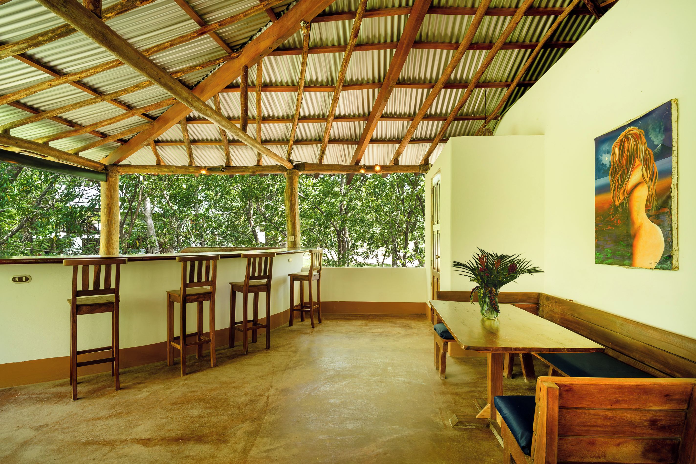 A covered outdoor seating area with wooden furniture, a painting on the wall, and a view of trees through the open sides.