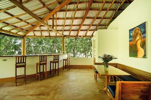 A covered outdoor seating area with wooden furniture, a painting on the wall, and a view of trees through the open sides.