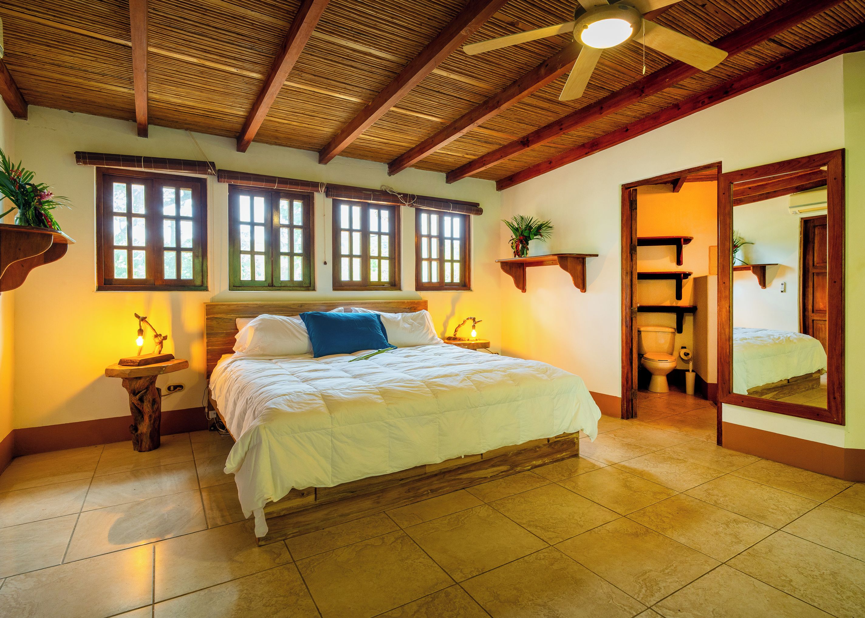 A bedroom with a wooden ceiling, a bed with white bedding, a nightstand with a lamp, and a bathroom visible through an open door.