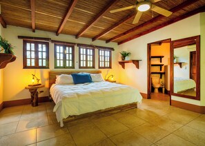 A bedroom with a wooden ceiling, a bed with white bedding, a nightstand with a lamp, and a bathroom visible through an open door. - Spacious 3BR, 3.5BA home just minutes from Playa Maderas (San Juan del Sur)