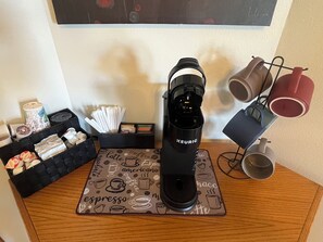 Eine Kaffeemaschine auf einem Holztisch mit Untertasse im Kaffeemotiv, einer Kapselkorb und einem Mughalter mit zwei Tassen.