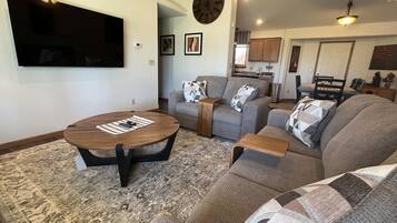 A living room with a flat-screen TV, a round coffee table, and two sofas with patterned throw pillows.