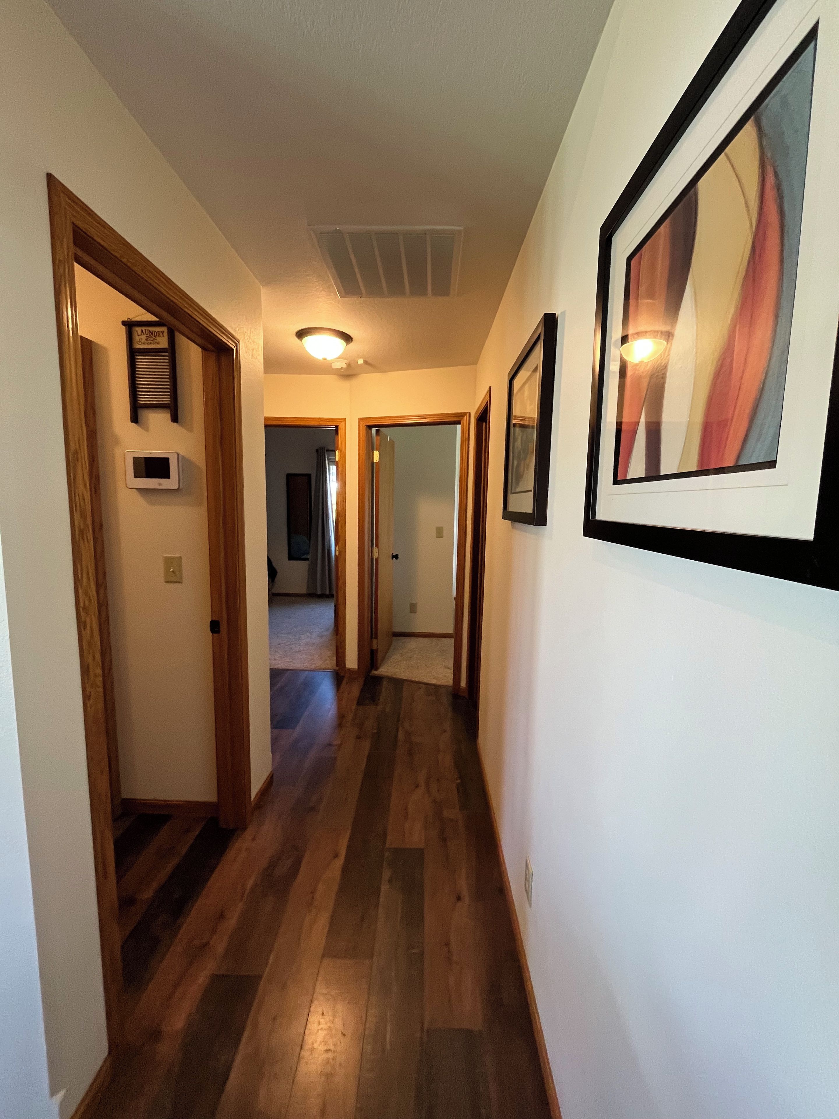 A hallway with wooden flooring, framed artwork on the walls, and a visible laundry sign.