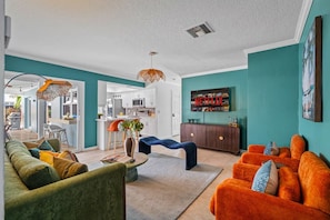 A modern living room with a sofa, armchair, and ottoman, a kitchen in the background, and a television displaying 'NETFLIX' on the wall. - Heated pool, hot tub, sauna, golf, theater, lounge (West Palm Beach)