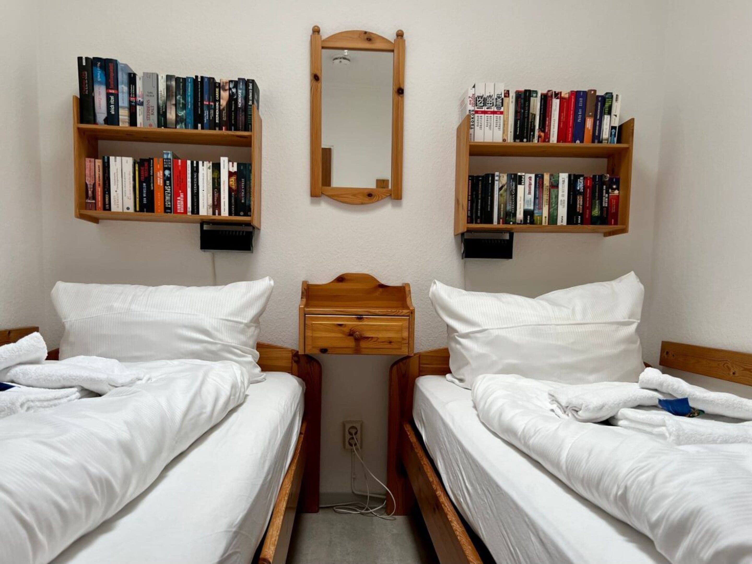 A bedroom with two single beds, a wooden nightstand, a mirror, and two bookshelves filled with books.