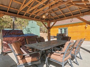 An outdoor patio with a covered seating area, a grill, and a wooden cabin in the background. - New! Bass Lake Escape with hot tub/game room/steps to the lake! (Bass Lake)