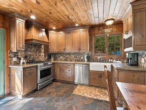 A kitchen with wooden cabinets, a stone backsplash, a built-in oven, a dishwasher, and a window with a view of greenery. - New! Bass Lake Escape with hot tub/game room/steps to the lake! (Bass Lake)