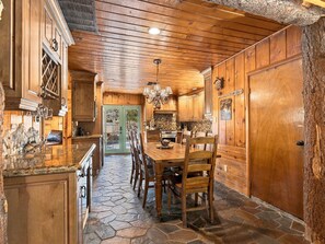 A rustic kitchen with wooden cabinets, a stone floor, and a dining area with chairs. - New! Bass Lake Escape with hot tub/game room/steps to the lake! (Bass Lake)