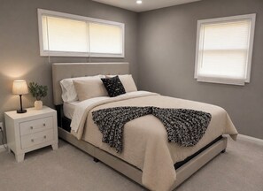 Ein Schlafzimmer mit einem Bett, einem Nachttisch, einer Lampe und zwei Fenstern.