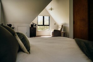 Ein Schlafzimmer mit schräg steilender Decke, einem großen Fenster, einem Bett, einem Sessel und einem kleinen Tisch.