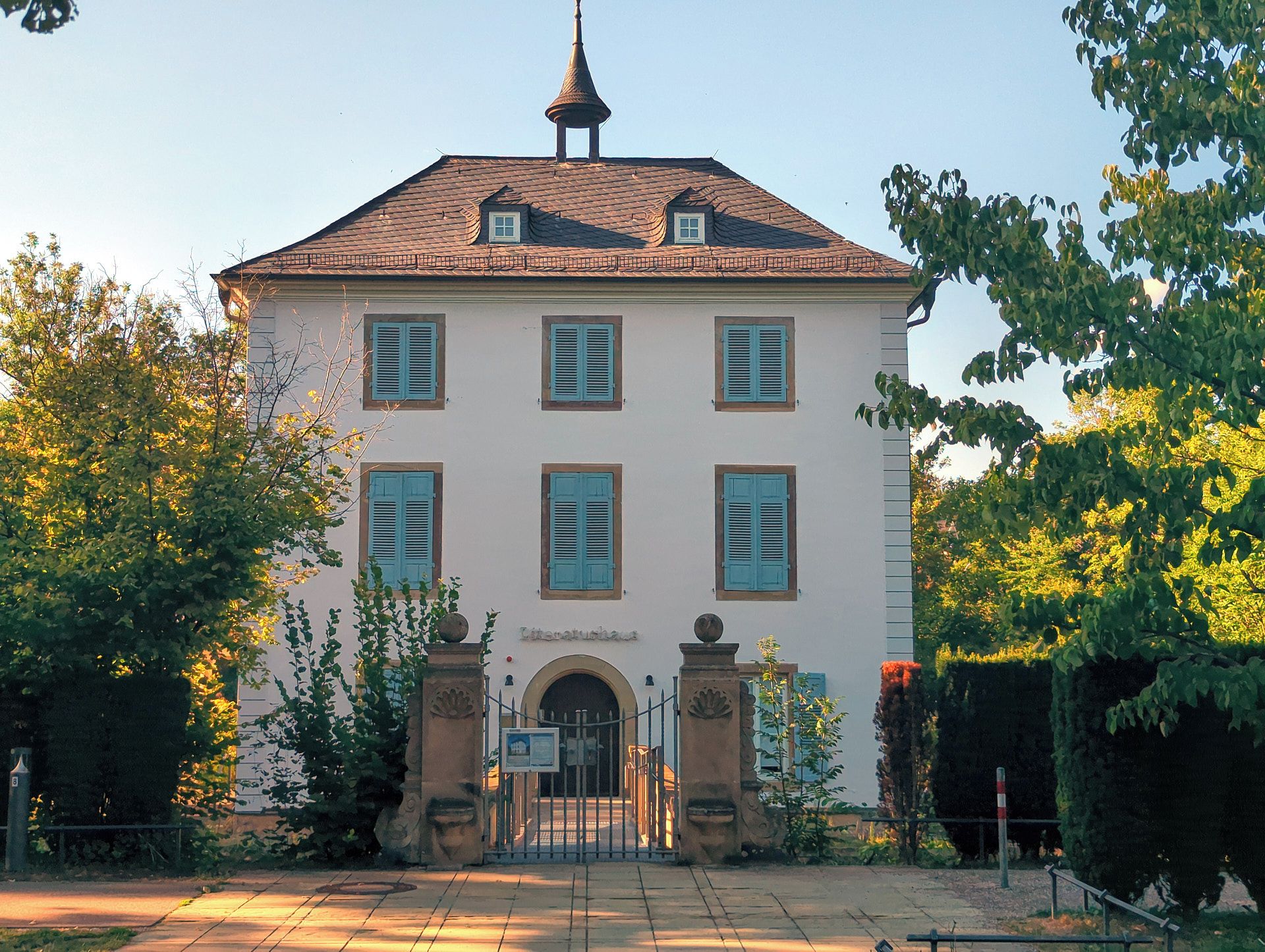 Ein zweistöckiges Gebäude mit einer symmetrischen Fassade, einem zentralen Eingang mit einer Tür und einem kleinen turmartigen Aufsatz auf dem Dach.