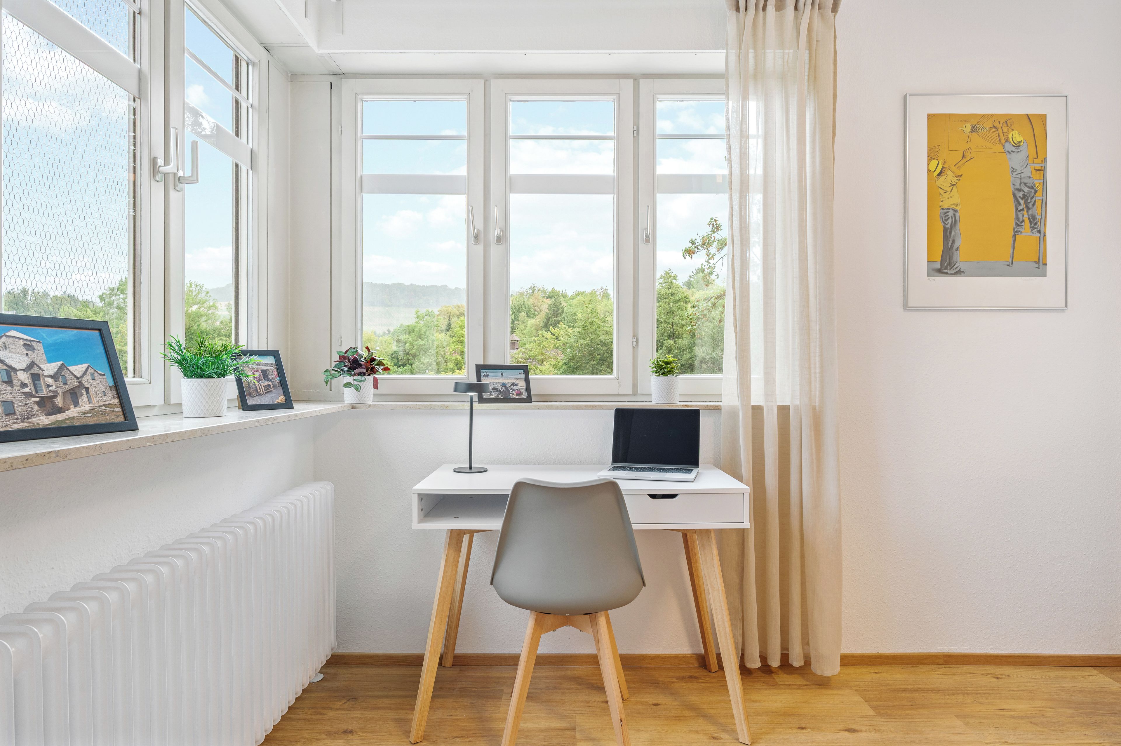 Ein Zimmer mit Schreibtisch, Stuhl und einem Fenster mit Vorhängen. Auf dem Fensterbrett befinden sich gerahmte Bilder und darunter ein Heizkörper.