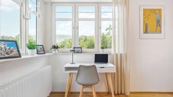 Ein Zimmer mit Schreibtisch, Stuhl und einem Fenster mit Vorhängen. Auf dem Fensterbrett befinden sich gerahmte Bilder und darunter ein Heizkörper.
