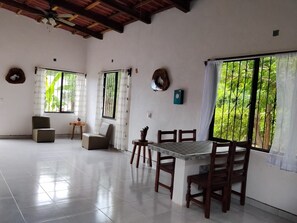 A spacious room with a tiled floor, a dining table with chairs, a ceiling fan, and large windows with curtains.