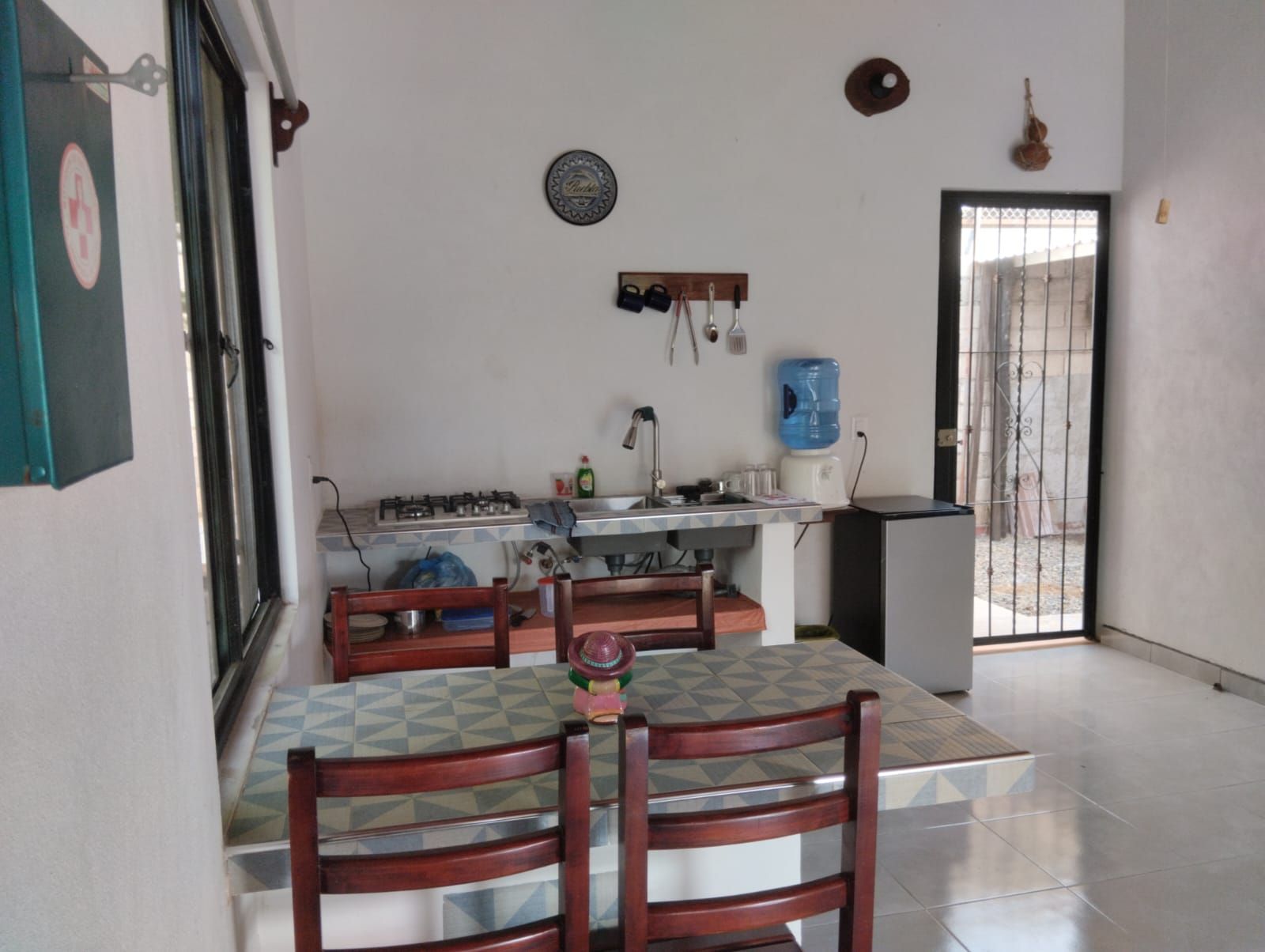A kitchen with a gas stove, sink, and a water dispenser. There is a dining table with chairs and a decorative plate on the wall.