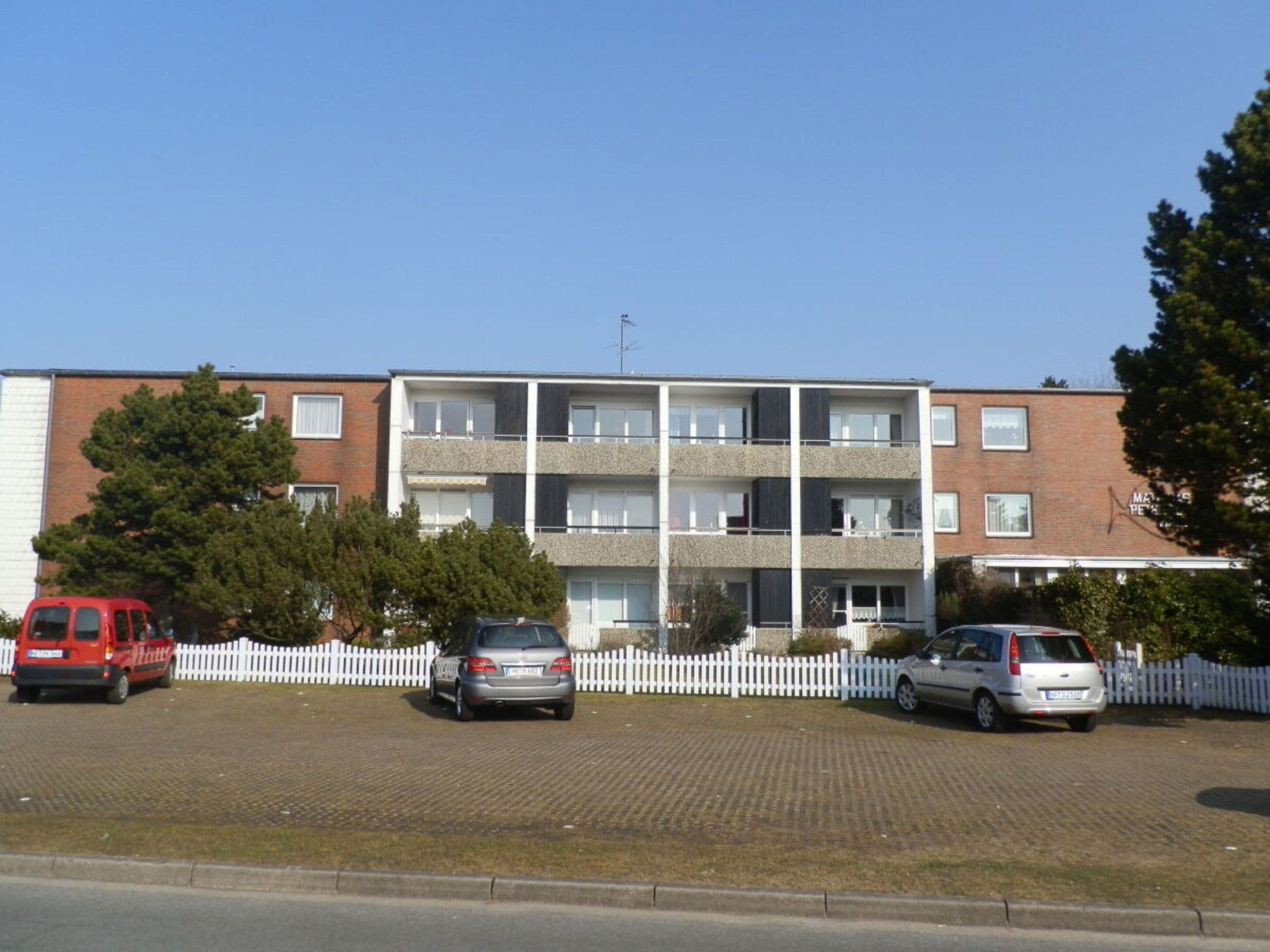 A multi-story residential building with a white picket fence, parked cars, and a clear sky.