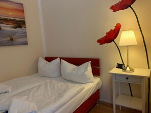A bed with white bedding and red headboard, a bedside table with a lamp, and a wall-mounted painting of a sunset. - Holiday Apartment With 2 Bedrooms (Wyk auf Foehr)