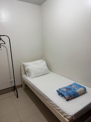 Un lit simple avec un matelas et des oreillers blancs, une couverture bleue à motifs et une armoire en métal noir, dans une pièce aux murs blancs et au sol carrelé.