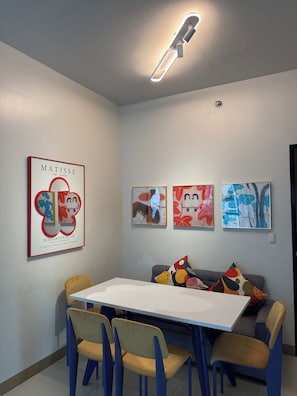 Un coin repas avec une table blanche et des chaises bleues, un canapé gris agrémenté de coussins colorés, et trois œuvres d’art encadrées accrochées au mur.