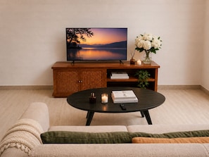 Een woonkamer met een flatscreen televisie op een houten kast, een ronde salontafel met boeken en kaarsen, en een bank met een plaid.