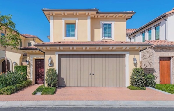 Una casa de dos pisos con fachada color beige, un gran portón de garaje y un camino de ladrillo que conduce a la entrada.