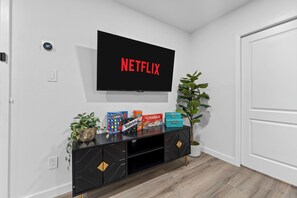 A modern living room with a flat-screen TV displaying the Netflix logo, a black entertainment unit with board games, and a potted plant. - 2-bedroom Duo apartment in marvelous Dallas with AC and fitness room (Dallas)